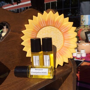 May include: Three small glass bottles with yellow liquid and black roll-on tops. The bottles are labeled "Movement (Constipation Relief)". The bottles are sitting on a wooden shelf with a yellow sunflower-shaped plate behind them.