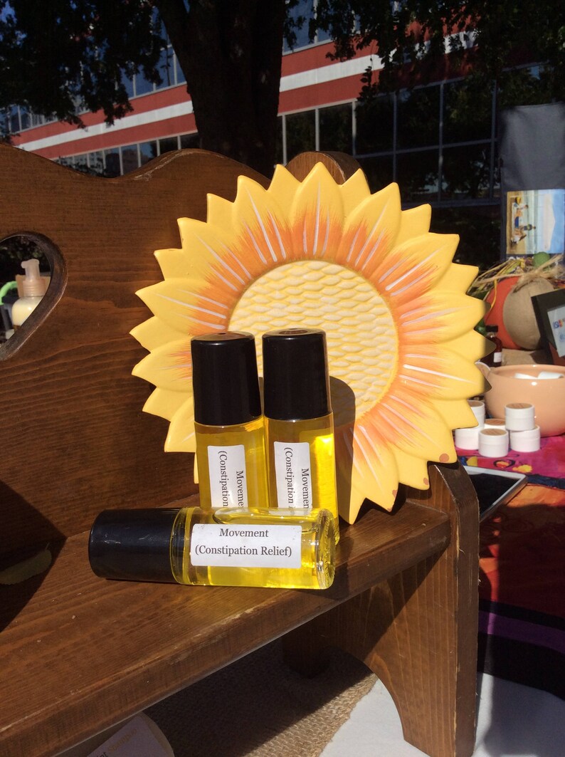 May include: Three small glass bottles with yellow liquid and black roll-on tops. The bottles are labeled "Movement (Constipation Relief)". The bottles are sitting on a wooden shelf with a yellow sunflower-shaped plate behind them.