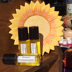 May include: Three amber glass roller bottles with black caps and labels that read "Movement (Constipation Relief)". The bottles are sitting on a wooden shelf with a yellow sunflower decoration behind them.