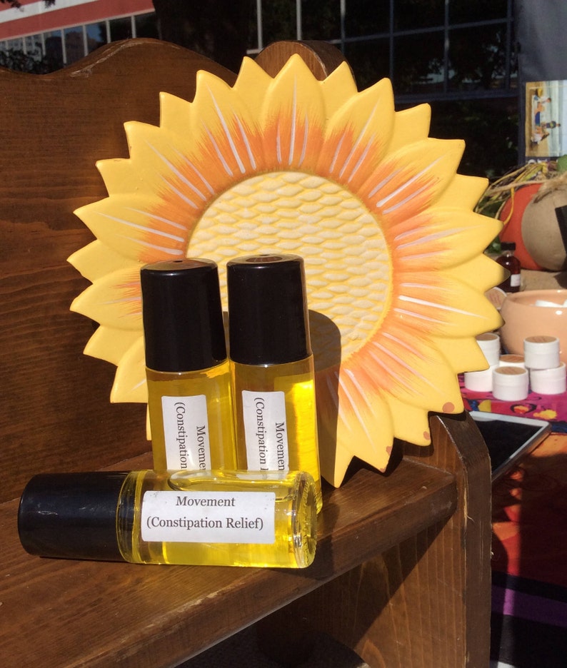 May include: Three amber glass roller bottles with black caps and labels that read "Movement (Constipation Relief)". The bottles are sitting on a wooden shelf with a yellow sunflower decoration behind them.