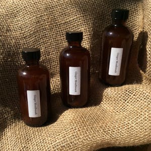May include: Three brown glass bottles with black caps and white labels that say "Ginger Mouthwash". The bottles are sitting on a brown burlap surface.