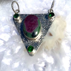 May include: A triangular silver pendant featuring a large oval ruby zoisite cabochon in the centre, accented by three small round green gemstones set at the corners. The silver frame has an intricate filigree pattern and two top loops.