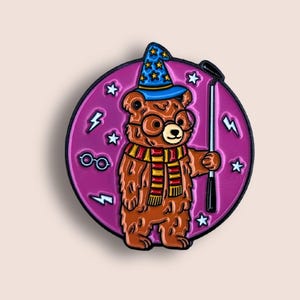 May include: A circular enamel pin featuring a brown bear wearing a blue wizard hat with yellow stars, round glasses, and a red and yellow striped scarf. The bear holds a black golf club against a purple background with white stars and lightning bolts.