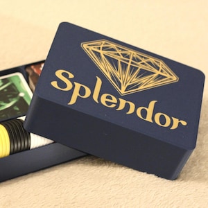 Könnte beinhalten: Eine dunkelblaue, rechteckige Spielaufbewahrungsbox mit einer goldenen Diamantgrafik und dem Schriftzug Splendor auf dem Deckel. Die geöffnete Box zeigt Innenfächer mit einem Kartenspiel mit dem Wort Splendor sowie Stapeln von gelben und schwarzen Spielsteinen.