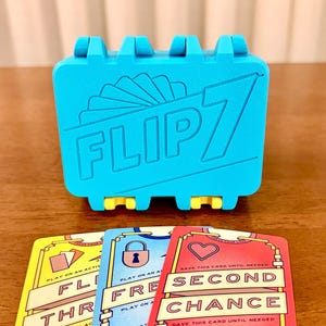 Op de afbeelding: Een felcyaan plastic kaartenhouder met de in reliëf aangebrachte tekst FLIP 7 en een icoon van een waaier kaarten. Drie spelkaarten liggen ervoor, gekleurd in geel, blauw en rood, met teksten zoals SECOND CHANCE, FREE en THR, en diverse iconen.