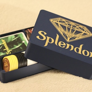 Può includere: Una scatola portaoggetti per giochi blu scuro con un grafico a diamante dorato e il testo Splendor stampato sul coperchio. La scatola aperta contiene una pila di carte da gioco rettangolari e una fila di gettoni rotondi gialli, neri e bianchi.
