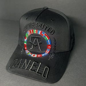 May include: A black baseball cap featuring the word UNDISPUTED embroidered in black above a circular emblem of various international flags surrounding a black CA logo. The word CANELO is embroidered in black on the curved brim of the cap.