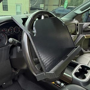 May include: A black, textured steering wheel tray mounted onto a vehicle steering wheel. The tray features a flat, rectangular surface with a raised bottom edge to hold items, designed to fit securely over the top portion of the steering wheel.