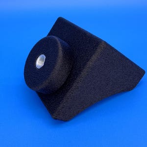 May include: A black, textured, triangular-shaped mounting bracket featuring a circular protrusion on one side. The center of the circular section contains a silver-colored, threaded metal insert designed for a screw or bolt attachment.