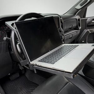 May include: A black steering wheel desk tray mounted onto a vehicle steering wheel, supporting a silver laptop computer. The tray is constructed from a dark, textured material and provides a flat surface for the laptop within the car interior.