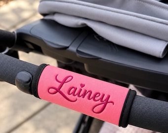 May include: A bright pink stroller handlebar cover featuring the name Lainey embroidered in a dark pink cursive script with a small heart detail above the letter i. The cover is secured around a black foam stroller handle with black trim.