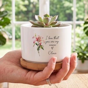 Könnte beinhalten: Ein zylindrischer Übertopf aus weißer Keramik mit Bambussockel, der eine grüne Sukkulente enthält. Der Übertopf ist mit einer rosa Lilienillustration und dem schwarzen Text bedruckt: I love that, you are my mama, einem Herzsymbol und dem Namen Olivia.