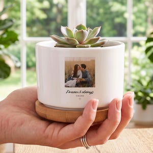 May include: A white ceramic cylindrical planter with a light wood base, containing a green succulent. The front features a square photograph of two people in a field, with the text "Forever & always" and the date "11.05.2025" printed below.