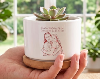 May include: A white ceramic cylindrical planter with a bamboo base, containing a green succulent. The planter features a red line-art illustration of two people hugging and the text, No One Will Ever Be As Entertained By Us As Us.