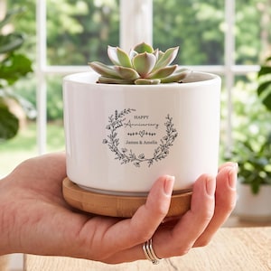 May include: A white ceramic cylindrical planter with a bamboo base, containing a green succulent. The front features a black printed floral wreath design with the text "HAPPY Anniversary" and "James & Amelia" centered inside the wreath.