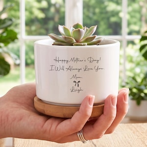 May include: A white ceramic cylindrical planter with a bamboo base, containing a green succulent. The planter features black printed text that reads: Happy Mother's Day! I Will Always Love You. Mom. followed by a small bow icon and the name Leigh.