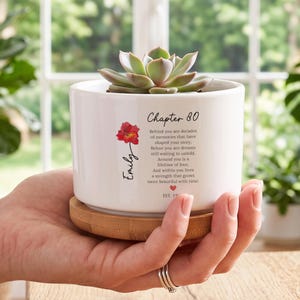 May include: A white ceramic planter with a bamboo base containing a succulent. The planter features a red flower graphic, the name Emily, and text reading Chapter 80, followed by a poem about memories, love, and strength, ending with a heart and EST. 19.