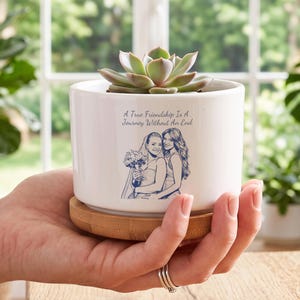 Peut inclure: Un pot de fleurs cylindrique en céramique blanche avec une base en bambou, contenant une plante succulente verte. L'avant présente une illustration au trait bleu de deux personnes, dont une en voile de mariée, avec le texte A True Friendship Is A Journey Without An End.