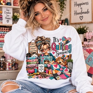 May include: A white long-sleeve sweatshirt featuring a graphic illustration of a person with a leopard print headband sewing at a machine. The design includes a cat, a dog, a colorful quilt, and the text It's Quilting Season and Quilting Fuels My Soul.