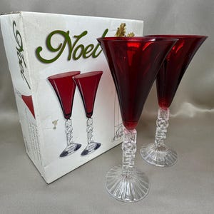 May include: Two red glass goblets with clear stems featuring the word NOEL embossed vertically. The base of each stem is fluted. The glasses are displayed next to a white cardboard box with the word NOEL printed in green script.