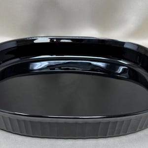 May include: An oval-shaped baking dish with a glossy black finish. The dish features a smooth, deep interior basin and a fluted exterior wall design with vertical ridges running along the entire perimeter of the vessel's outer edge.