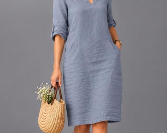 May include: A knee-length, slate blue dress with a V-neckline, three-quarter length sleeves with button tabs, and side pockets. The dress is paired with white sneakers and a woven straw handbag containing small white flowers.