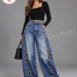 Op de afbeelding: Een zwart shirt met lange mouwen en een vierkante halslijn, gecombineerd met een wijde blauwe spijkerbroek met goudkleurige metalen ringen langs de buitenste naden. De outfit bevat een kleine zwarte handtas en witte sneakers, met het DreamClothingHub-logo.