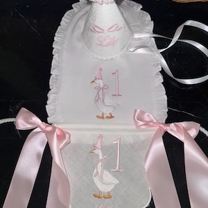 Könnte beinhalten: Weißes Lätzchen- und Banner-Set mit rosa Akzenten, entworfen für eine erste Geburtstagsfeier. Mit gestickter Gans mit Partyhut, der Zahl 1 und dem Namen "Lily". Inklusive passendem Partyhut mit rosa Bommel.