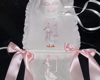 May include: White bib and banner set with pink accents, designed for a first birthday celebration. Features embroidered goose wearing a party hat, the number 1, and the name "Lily". Includes a matching party hat with a pink pom-pom.