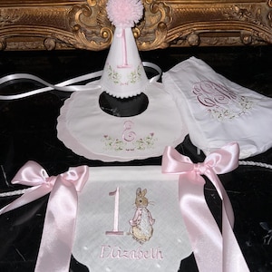 May include: A set of baby's first birthday accessories. Includes a white party hat with a pink pom-pom, a bib, and a banner featuring Peter Rabbit. All items are white with pink embroidery and accents.