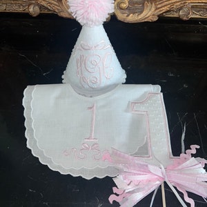 May include: A white embroidered bib with a pink number one and a pink bow. A pink pom-pom hat with the letter "H" embroidered on it is on top of the bib. A pink and white streamer is in the background.