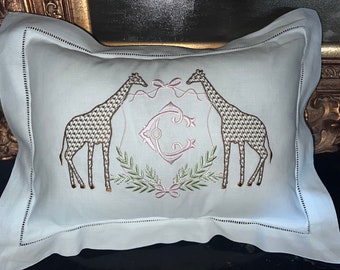 Może przedstawiać: Biała poduszka z jasnoniebieskim tłem, na której wyszyto żyrafy, różowy monogram i zielone liście.