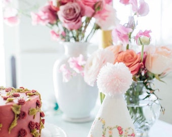May include: A white embroidered party hat with a pink pom-pom on top. The hat says "One" in colorful embroidery. A pink and white floral arrangement is in the background.
