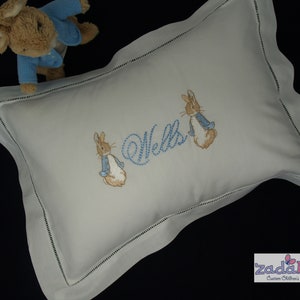 May include: A white rectangular pillow with a scalloped edge and embroidered details. The pillow features two bunnies and the name "Wells" in blue thread. A stuffed bunny wearing a blue jacket is in the background.