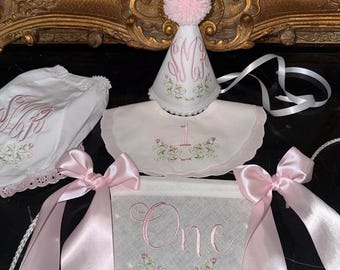 May include: A white party set featuring a cone-shaped hat with a pink pom-pom, a scalloped bib, and a pennant banner. Each piece is embroidered with pink monograms, green floral vines, and the word One in pink script with long pink satin ribbons.
