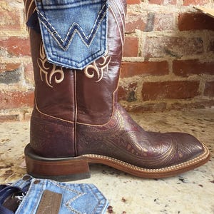 May include: A brown leather cowboy boot featuring a square toe, intricate gold paisley patterns on the foot, and decorative embroidery on the shaft. A blue denim pocket with a white W stitch is attached to the side, alongside a matching denim piece.