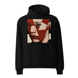 May include: A black hooded sweatshirt featuring a large square graphic on the chest. The graphic displays a stylized, fragmented portrait of a face in shades of deep red, off-white, and black, with a distressed, textured appearance throughout.