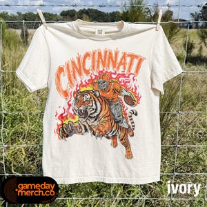 Peut inclure: Un t-shirt à manches courtes couleur ivoire avec une impression graphique représentant un joueur de football américain chevauchant un tigre entouré de flammes orange. Le mot CINCINNATI est imprimé en grandes lettres orange au-dessus.