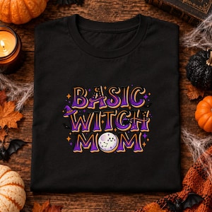 Può includere: Una t-shirt nera con il testo "BASIC WITCH MOM" in un carattere viola e arancione. La lettera "O" di "MOM" è sostituita da una luna bianca sorridente con un grafico a costellazione. Piccoli cappelli da strega viola e stelle arancioni circondano il testo.