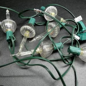 May include: A string of decorative holiday lights featuring clear, ribbed, bell-shaped glass covers over each bulb. The lights are connected by a dark green electrical cord and include a green plastic plug at the end of the wire strand.