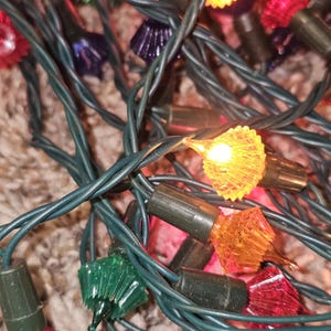 May include: A string of holiday lights featuring multi-colored, faceted plastic bulbs in red, green, orange, and purple. The bulbs are attached to dark green insulated wire, with some bulbs illuminated in a warm yellow glow.