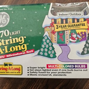 May include: A GE String-A-Long 70 light miniature set in a green box with a 32 ft length. The packaging features a winter house illustration, a 3 year guarantee, and multi-colored bulbs for indoor or outdoor use. The box includes safety information.