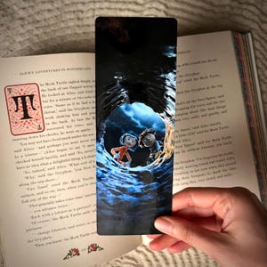 May include: A rectangular bookmark featuring a glossy, dark blue illustration of a tunnel looking out toward two animated characters, one with blue hair in an orange top and one with brown hair, set against an open book page of Alice's Adventures.