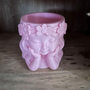 May include: A light pink planter shaped like a person's head with closed eyes and hands resting under the chin. The figure features wavy hair and a crown of sculpted flowers encircling the top rim, creating a smooth, matte finish throughout.