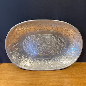 May include: An oval-shaped silver-toned metal tray featuring an embossed central floral motif surrounded by a diamond-patterned border. The outer rim is decorated with a repeating pattern of stylised tulip-like shapes and zigzag lines.