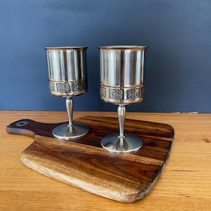 May include: Two silver-toned metal goblets featuring a smooth upper bowl and a decorative band with an embossed floral pattern around the middle. Each goblet has a slender stem and a circular base, providing a stable foundation for the vessel.