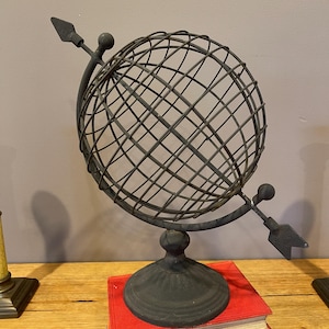 May include: A dark grey metal globe sculpture constructed from a wire grid frame. The globe is mounted on a circular pedestal base and features a decorative arrow passing through the centre, with a pointed tip and a fletched tail end.