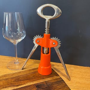 May include: A bright orange winged corkscrew with a metallic silver handle and gear mechanism, positioned next to a clear glass wine goblet. The corkscrew features a central vertical shaft and two downward-extending metal arms for leverage.