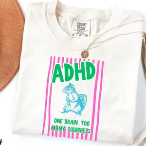 May include: An ivory t-shirt featuring a graphic print with the text ADHD in green, a blue squirrel illustration, and the text ONE BRAIN. TOO MANY SQUIRRELS. in green, all framed by vertical pink stripes. The shirt has a Comfort Colors brand label.