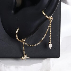 May include: A gold-toned double hoop earring set connected by a delicate chain. The lower hoop features a small star-shaped charm with embedded stones, while the upper hoop connects to a dangling marquise-shaped charm with a single stone accent.